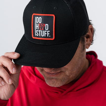 Man in red hoodie tipping the brim of the Do Hard Stuff Natural State hat with red and black patch.