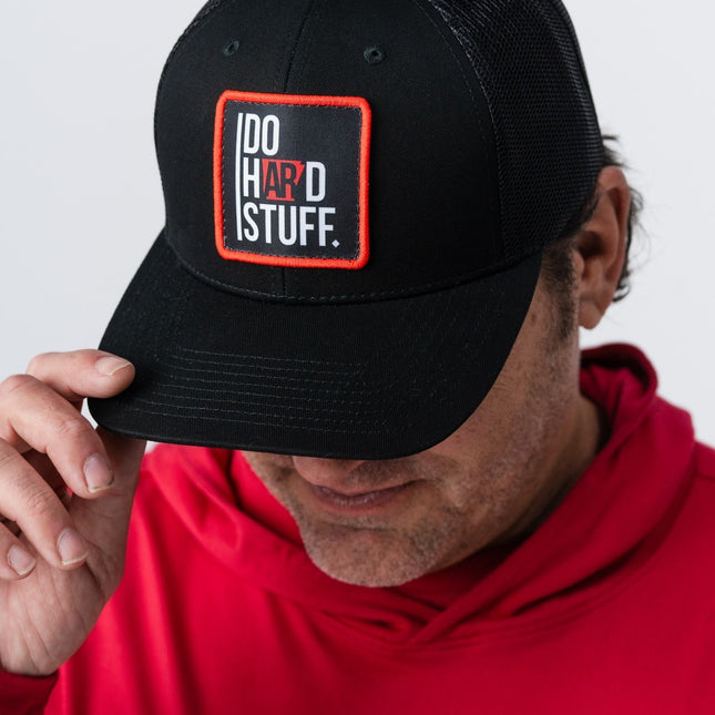 Man in red hoodie tipping the brim of the Do Hard Stuff Natural State hat with red and black patch.