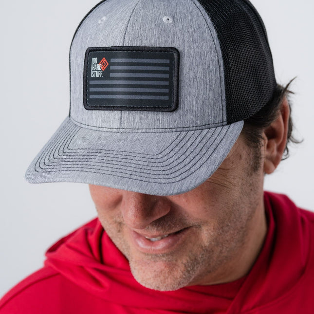 Do Hard Stuff Patriot Hat in close-up with gray front and black mesh, worn by man in red hoodie.