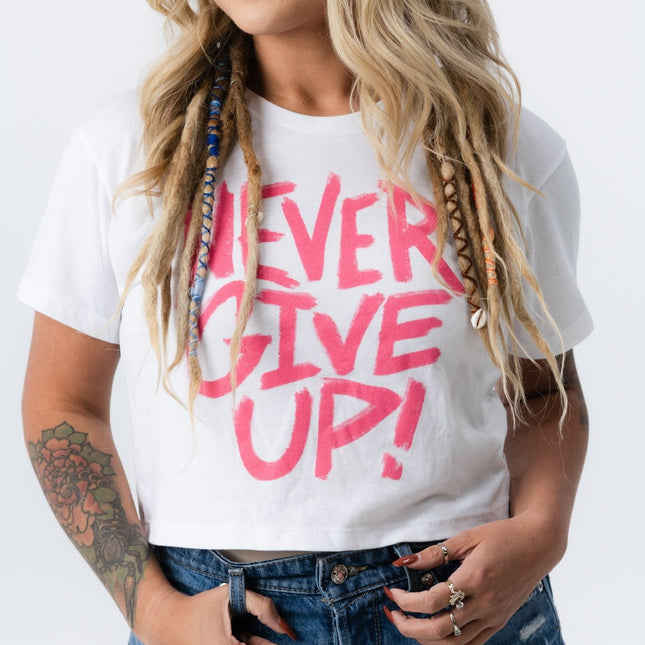 Side view of woman in pink and white "Never Give Up" crop tee with hands in jean pockets.