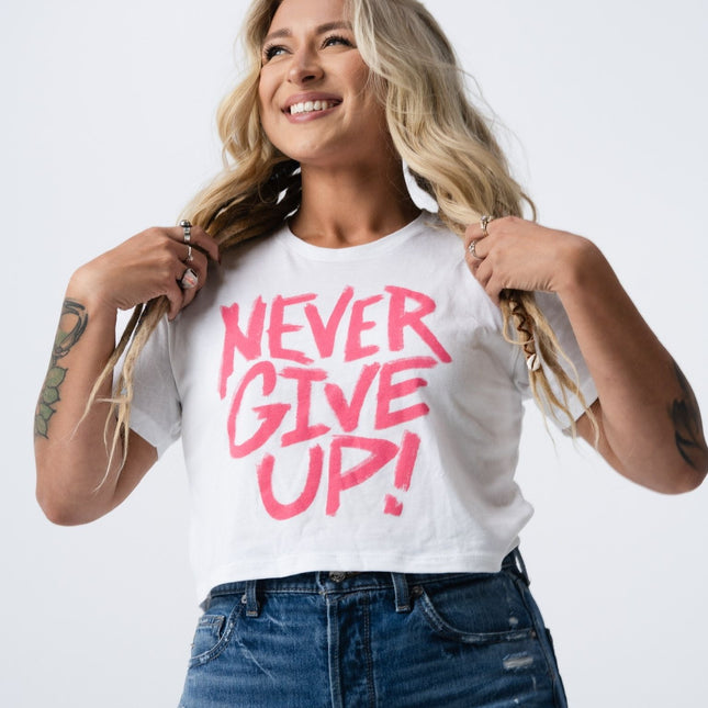 Model looking upward and smiling in the white crop tee with bold pink "Never Give Up" lettering.