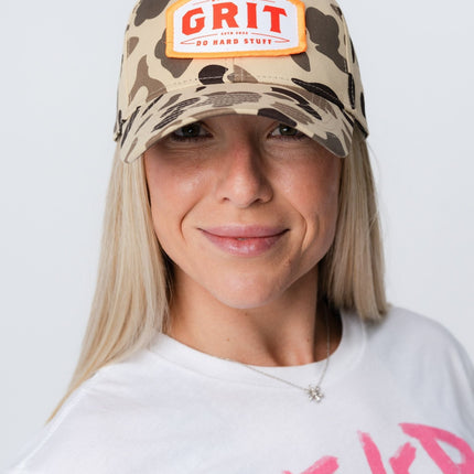 Woman wearing Grit Hat in vintage camo with bold orange patch and white lettering, front close-up