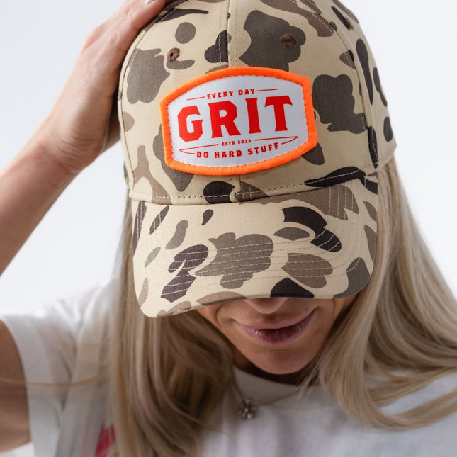 Woman adjusting top of vintage camo Grit Hat with vibrant orange and white patch