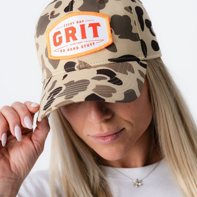 Side angle of woman tipping Grit Hat in vintage camo, showing detailed camo pattern and patch