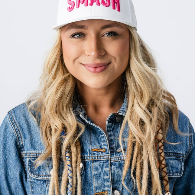 Woman in jean jacket tilting the brim of her pink SMASH hat, smiling in a side-profile pose.
